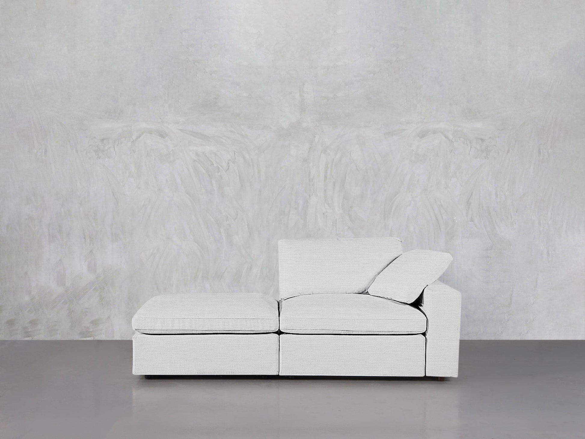 A modern 2-Seat Modular Lounger Loveseat in light gray with one armrest and loose cushions sits against a textured, light gray wall on a smooth, gray floor.