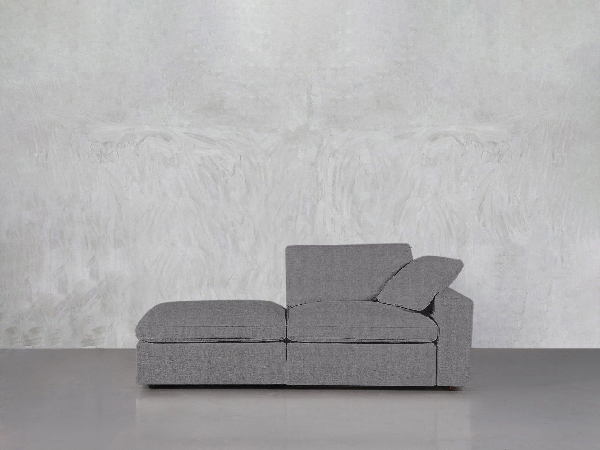 The 2-Seat Modular Lounger Loveseat, featuring clean lines and an angled cushion, is set on a smooth light gray floor against a textured white and gray abstract wall.