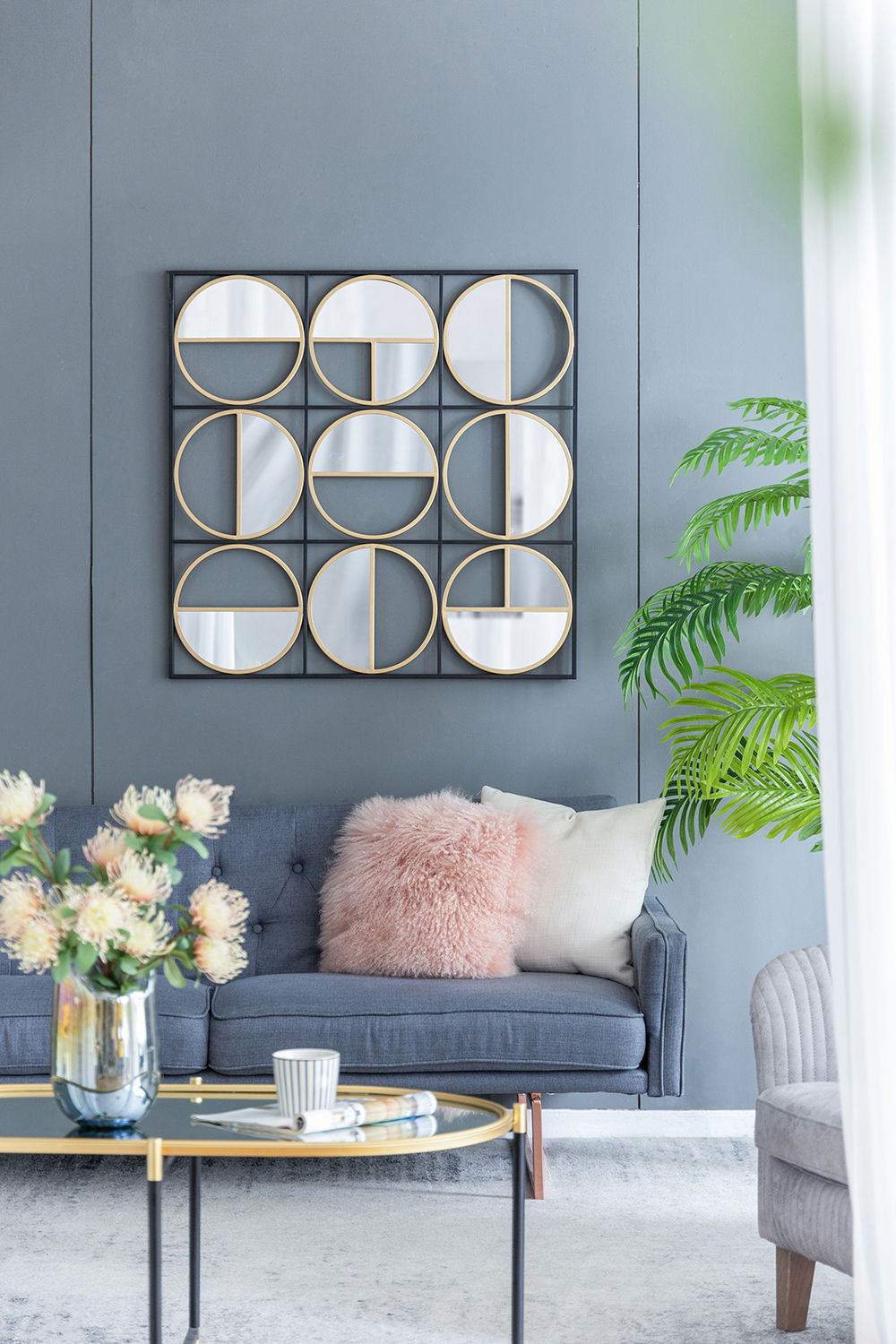 Modern living room with a gray sofa, pink and white pillows, a coffee table with flowers and cups, a potted plant, soft natural light from a window, and an Eclectic Styling Metal Beaded Wall Mirror with contemporary design.