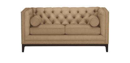 The Anderson Loveseat, in a beige hue with tufted upholstery, is showcased against a white background. It includes two cylindrical pillows and is adorned with nailhead trim along the edges, resting on dark wooden legs.