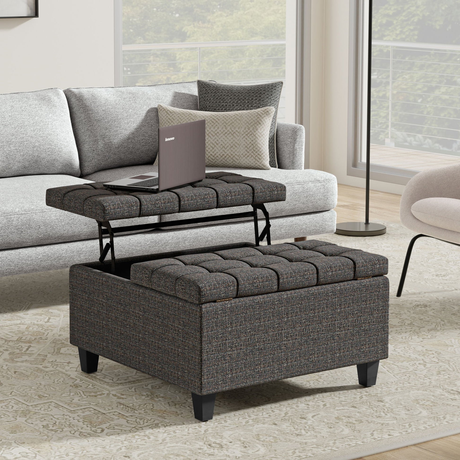 The Harrison Upholstered Coffee Table Storage Ottoman with a lift-top tray holding a laptop sits in front of a light gray sofa and next to a beige armchair in a modern living room with large windows.