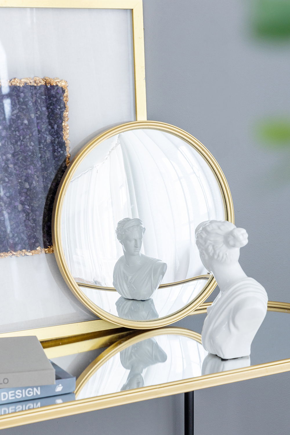 A white bust statue sits on a glass table next to books. Its reflection appears in the Round Mirror, Circle Mirror With Iron Frame, which leans against framed artwork on a gray wall.