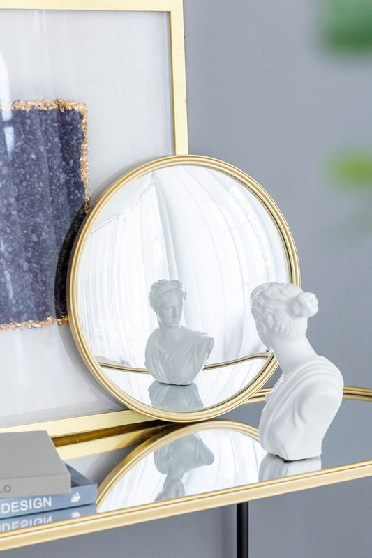 A white bust statue sits on a glass table next to books. Its reflection appears in the Round Mirror, Circle Mirror With Iron Frame, which leans against framed artwork on a gray wall.