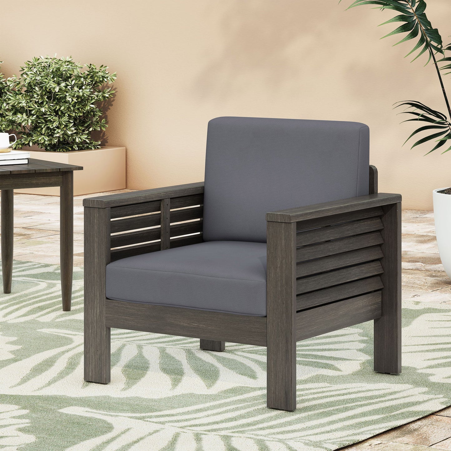 The Slat Design Club Chairs With Cushions, featuring a dark wooden frame and gray cushions, sit on a green and white leaf-patterned rug beside a table and potted plants on the patio.