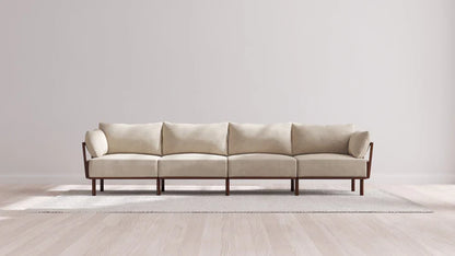 A minimalist room featuring the Indoor Jack 4-Seater Sofa in Balsa, characterized by its long design with wooden legs and arms, placed on a light-colored area rug. The wall and floor showcase shades of light, neutral tones, enhancing the modern, clean aesthetic.
