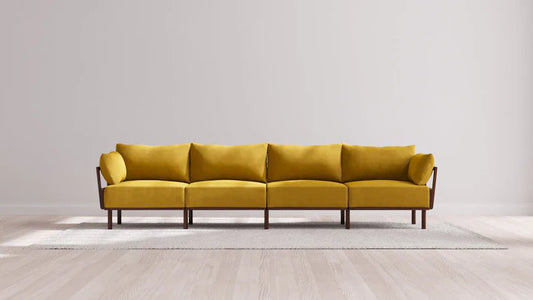 An Indoor Jack 4-Seater Sofa in Balsa, with a modern mustard yellow fabric and wooden legs, is centered in a minimalist room featuring light wooden flooring. The sofa rests on a neutral-colored area rug against an off-white wall.