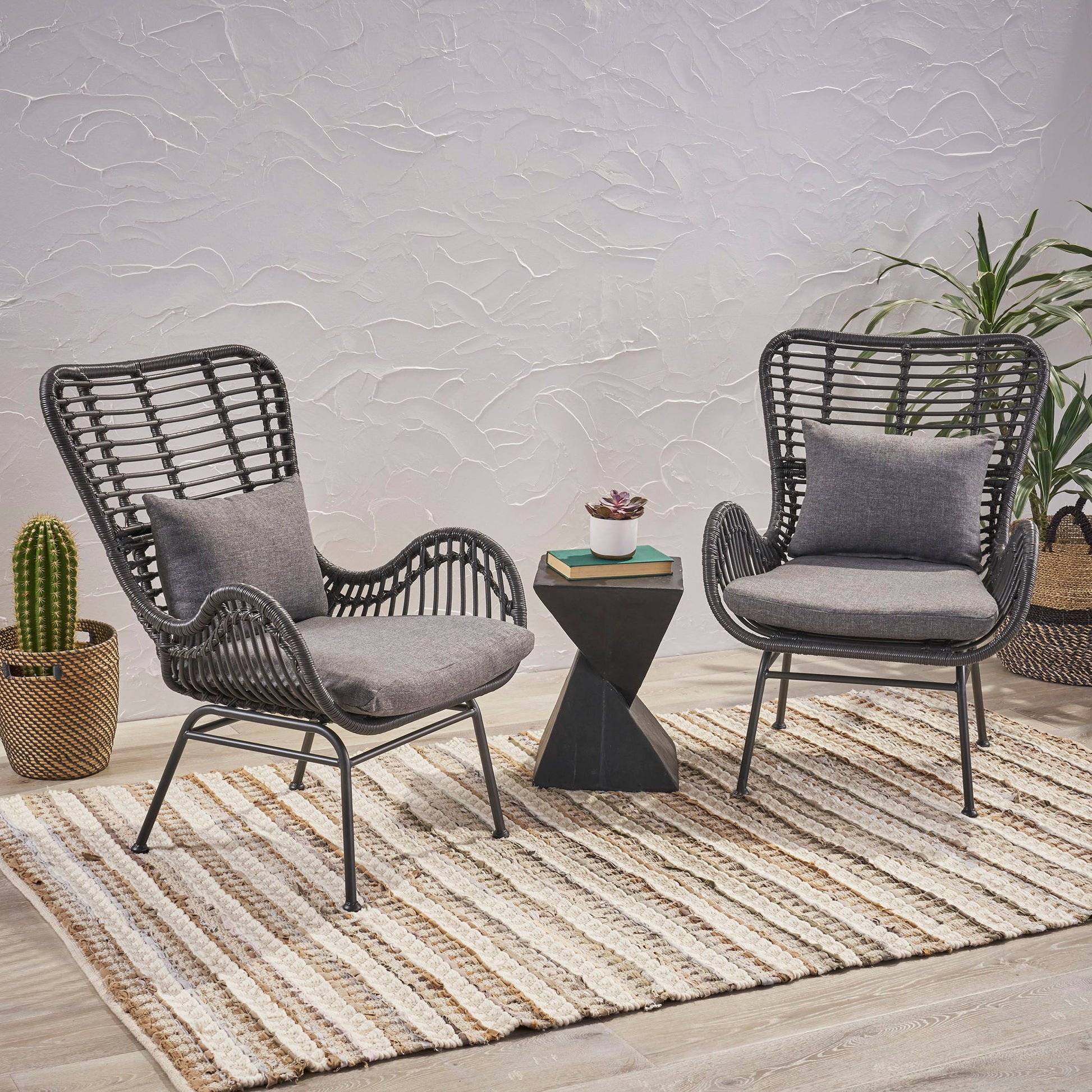 Two Modern Aesthetic Outdoor Rattan Accent Chairs with black frames and gray cushions sit on a striped rug, separated by a small black geometric table with a plant. In the background, potted cactus and palm plants are set against a textured white wall.
