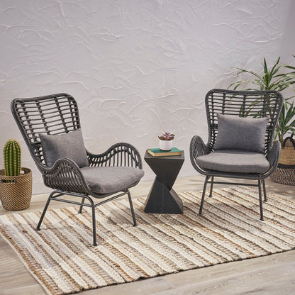 Two Modern Aesthetic Outdoor Rattan Accent Chairs with black frames and gray cushions sit on a striped rug, separated by a small black geometric table with a plant. In the background, potted cactus and palm plants are set against a textured white wall.