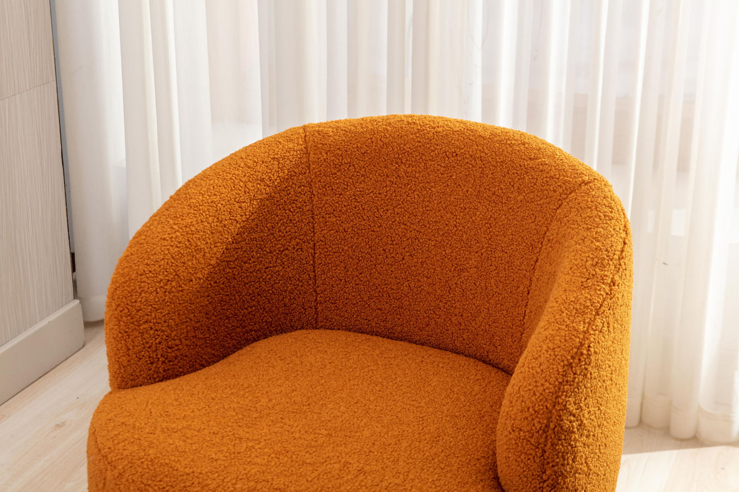 The Teddy Fabric Swivel Accent Armchair Barrel Chair with a powder-coated metal ring features modern curves and textured orange fabric, positioned in front of sheer white curtains on light wood flooring in a bright room.