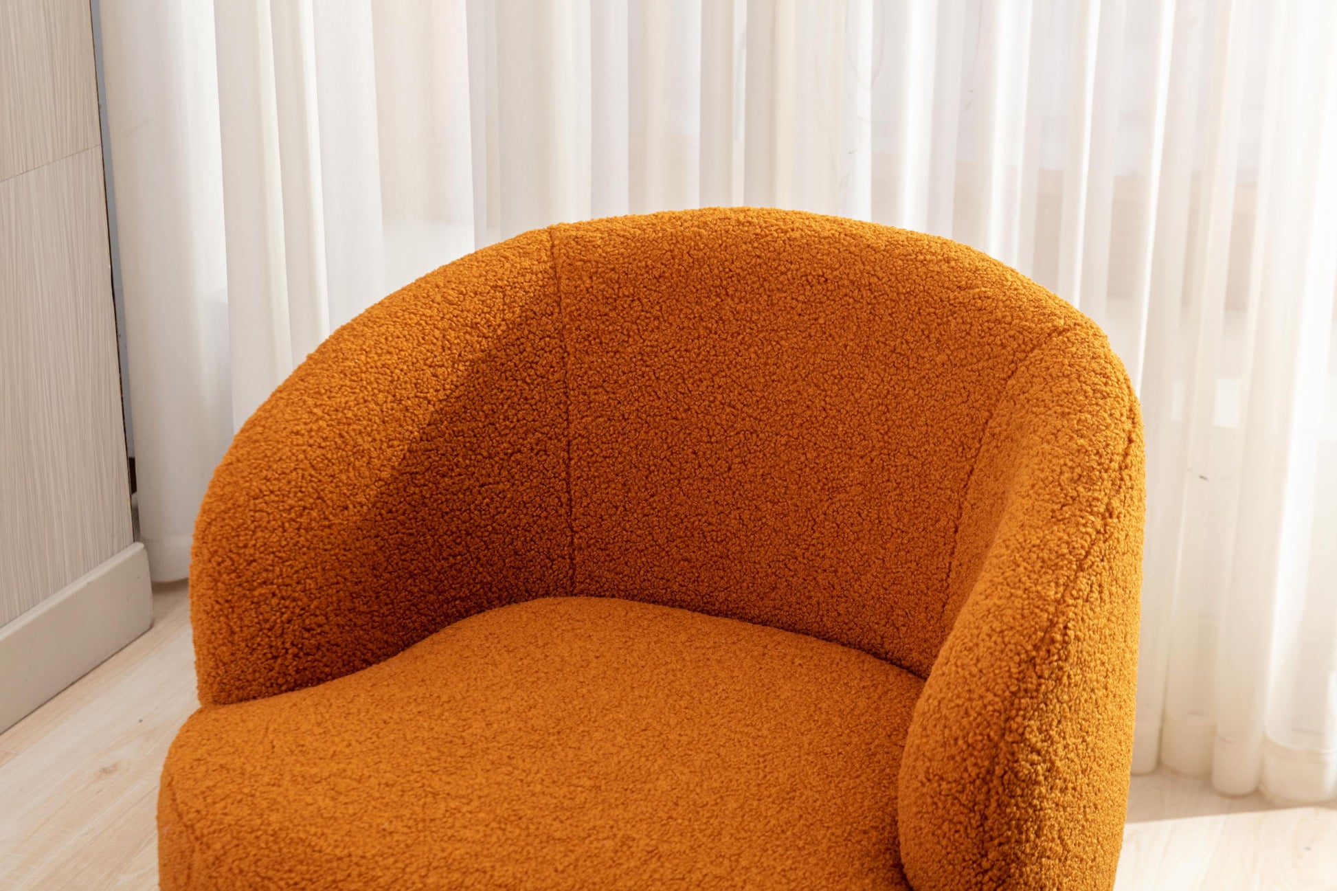 The Teddy Fabric Swivel Accent Armchair Barrel Chair with a powder-coated metal ring features modern curves and textured orange fabric, positioned in front of sheer white curtains on light wood flooring in a bright room.