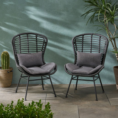 Two Calypso Outdoor Chairs with water-resistant gray cushions and iron frames sit side by side on a patio, surrounded by potted plants like cactus and palm, against a green textured wall with sunlight casting shadows on the scene.