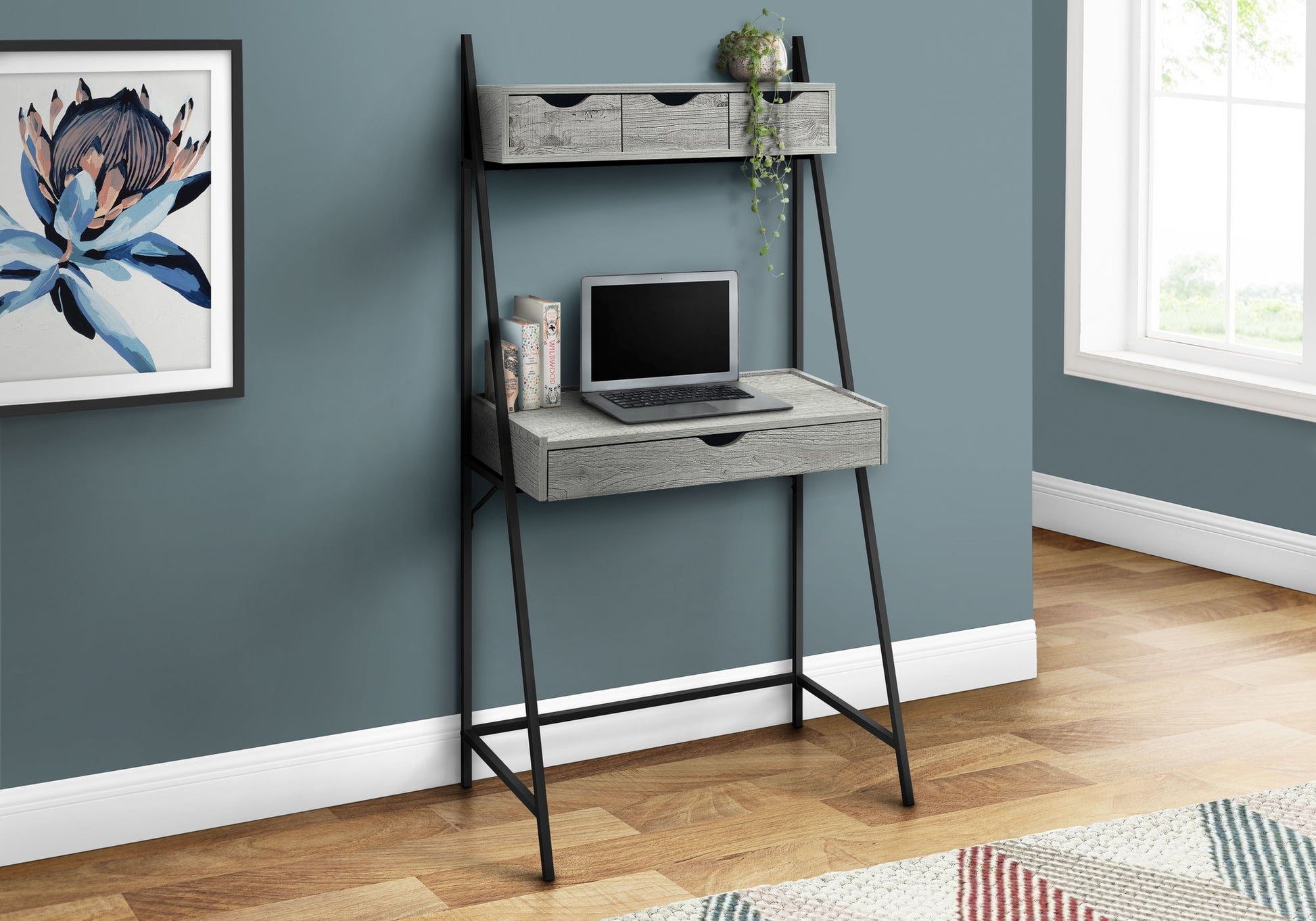 The Computer Desk For Home Office features black metal legs, gray shelves, three storage drawers, and an upper shelf with a potted plant. An open laptop sits on the desk against a blue wall near a framed flower print. Contemporary & modern style.