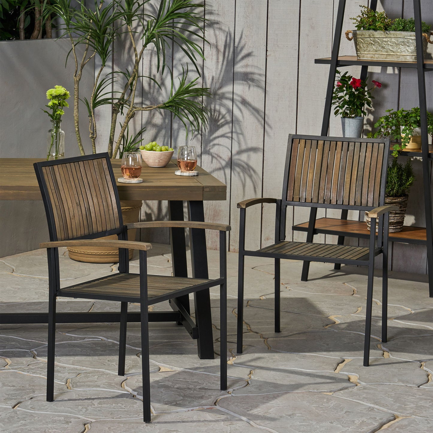 Two Vermilion Outdoor Dining Chairs with vertical slats and wooden arms stand on a stone patio next to an outdoor table topped with drinks and plants. In the background, potted plants and flowers are displayed on a shelf.