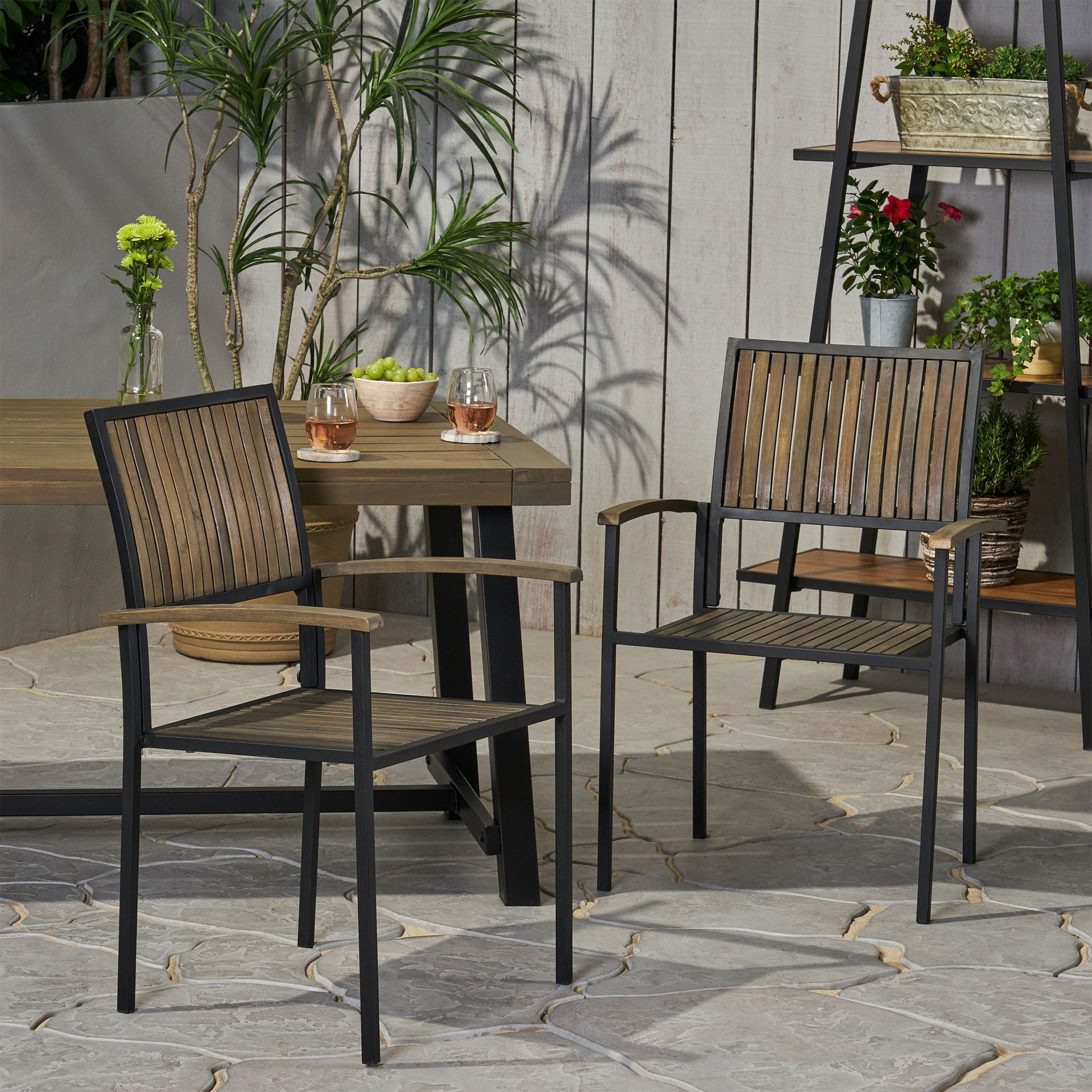 Two Vermilion Outdoor Dining Chairs with vertical slats and wooden arms stand on a stone patio next to an outdoor table topped with drinks and plants. In the background, potted plants and flowers are displayed on a shelf.
