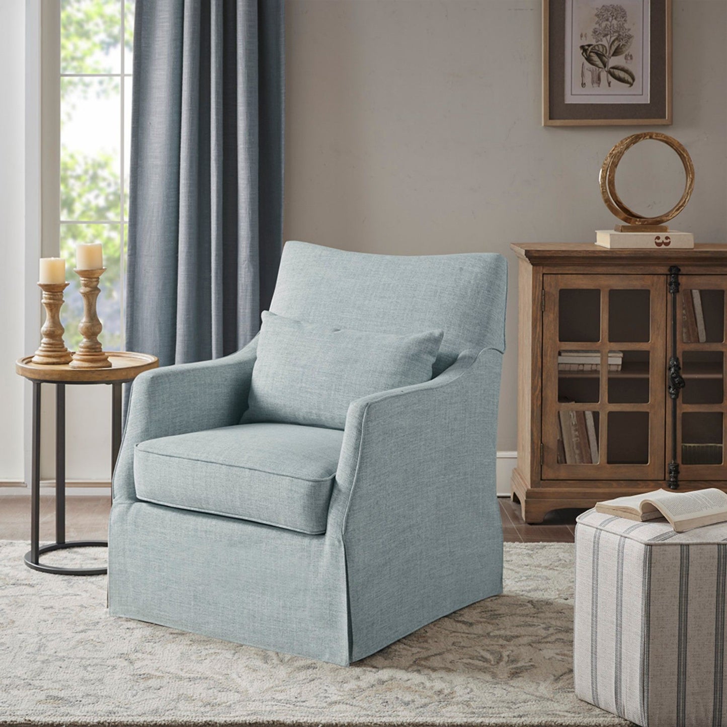 The Traditional Design London Skirted Swivel Chair in light blue, paired with a matching cushion, sits on a patterned rug beside a round side table with candles and a wooden decor cabinet. Sunlight streams in from the window.