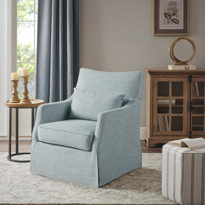 The Traditional Design London Skirted Swivel Chair in light blue, paired with a matching cushion, sits on a patterned rug beside a round side table with candles and a wooden decor cabinet. Sunlight streams in from the window.