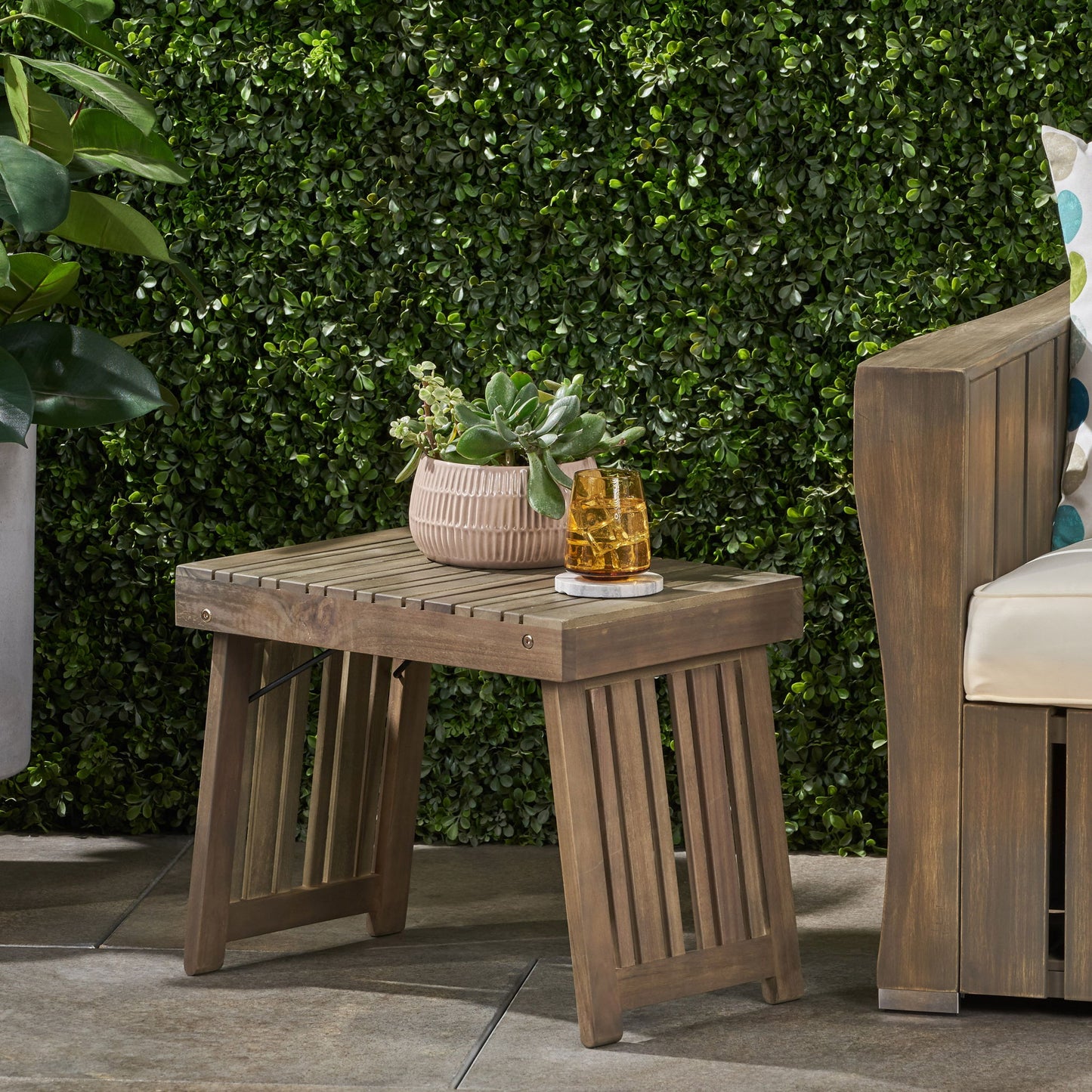 The Temptation Outdoor Foldable Side Table with Slat Design, topped with a potted succulent and two glasses, is set on a patio beside a wooden bench with white cushions, in front of a lush green hedge.