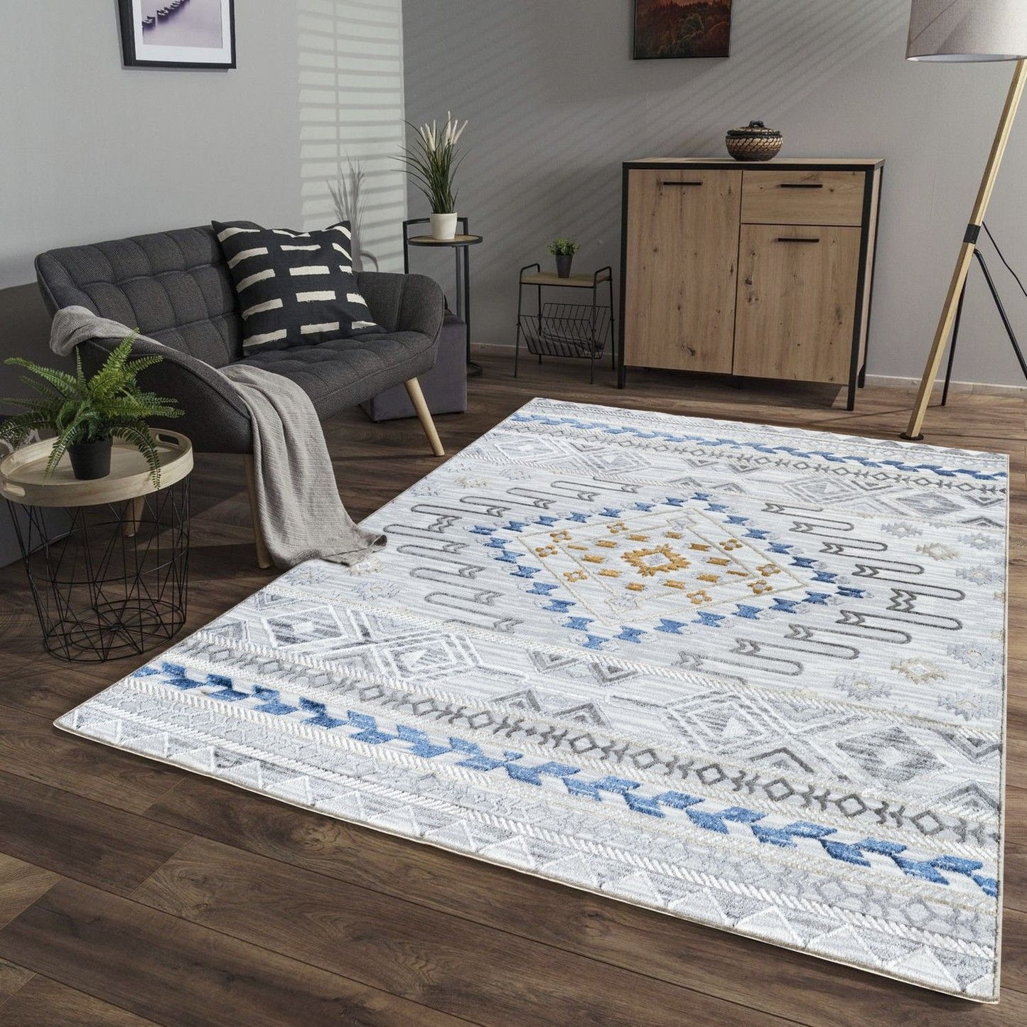 A modern living room with a gray sofa, striped pillow and blanket, wooden sideboard, large floor lamp, and the Legacy - GC_CAM8001 Area Rug featuring geometric patterns in white, blue, gold, and gray on a wooden floor.