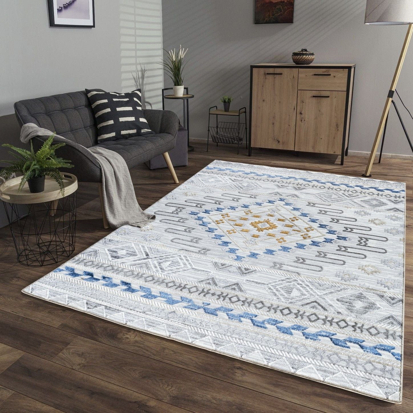 A modern living room with a gray sofa, striped pillow and blanket, wooden sideboard, large floor lamp, and the Legacy - GC_CAM8001 Area Rug featuring geometric patterns in white, blue, gold, and gray on a wooden floor.