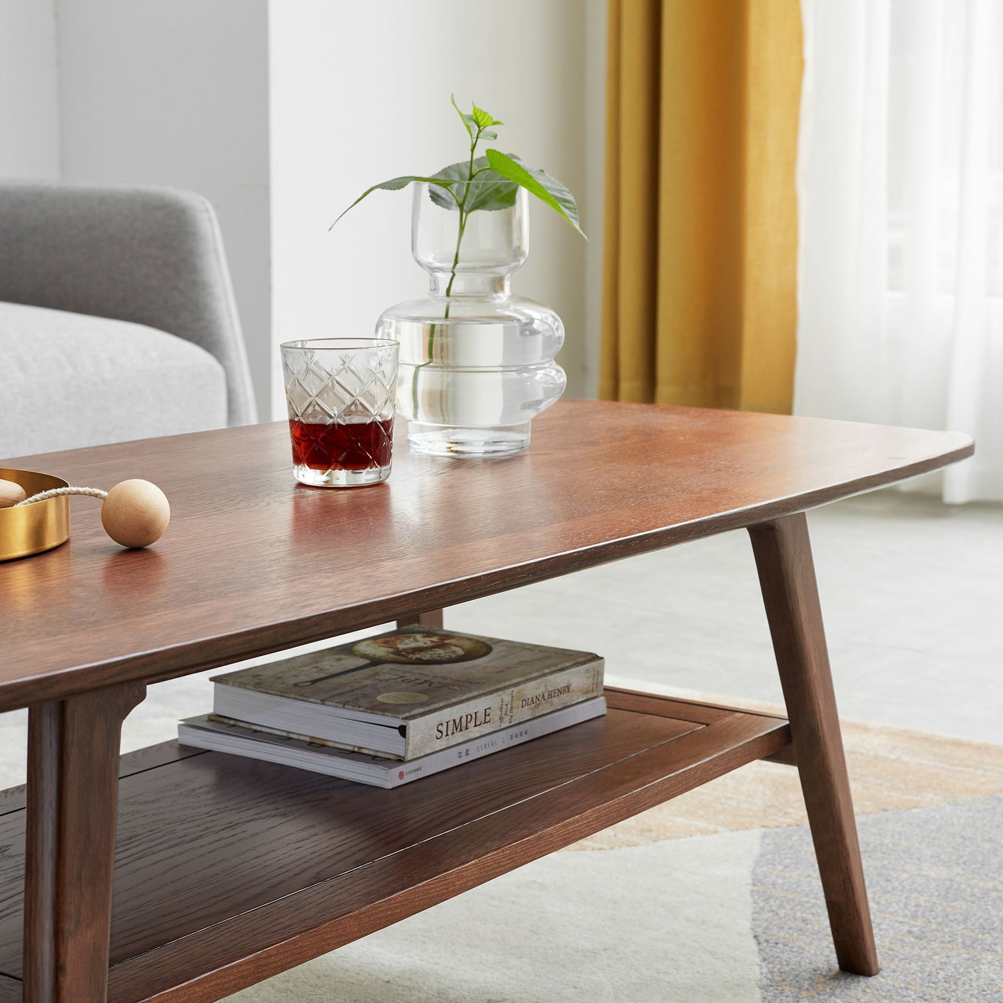 The Low Coffee Table With Storage Shelf features a glass of dark liquid, a clear vase with a green plant, a golden bowl with a wooden spoon, and books on the shelf below, all set in a bright living room.