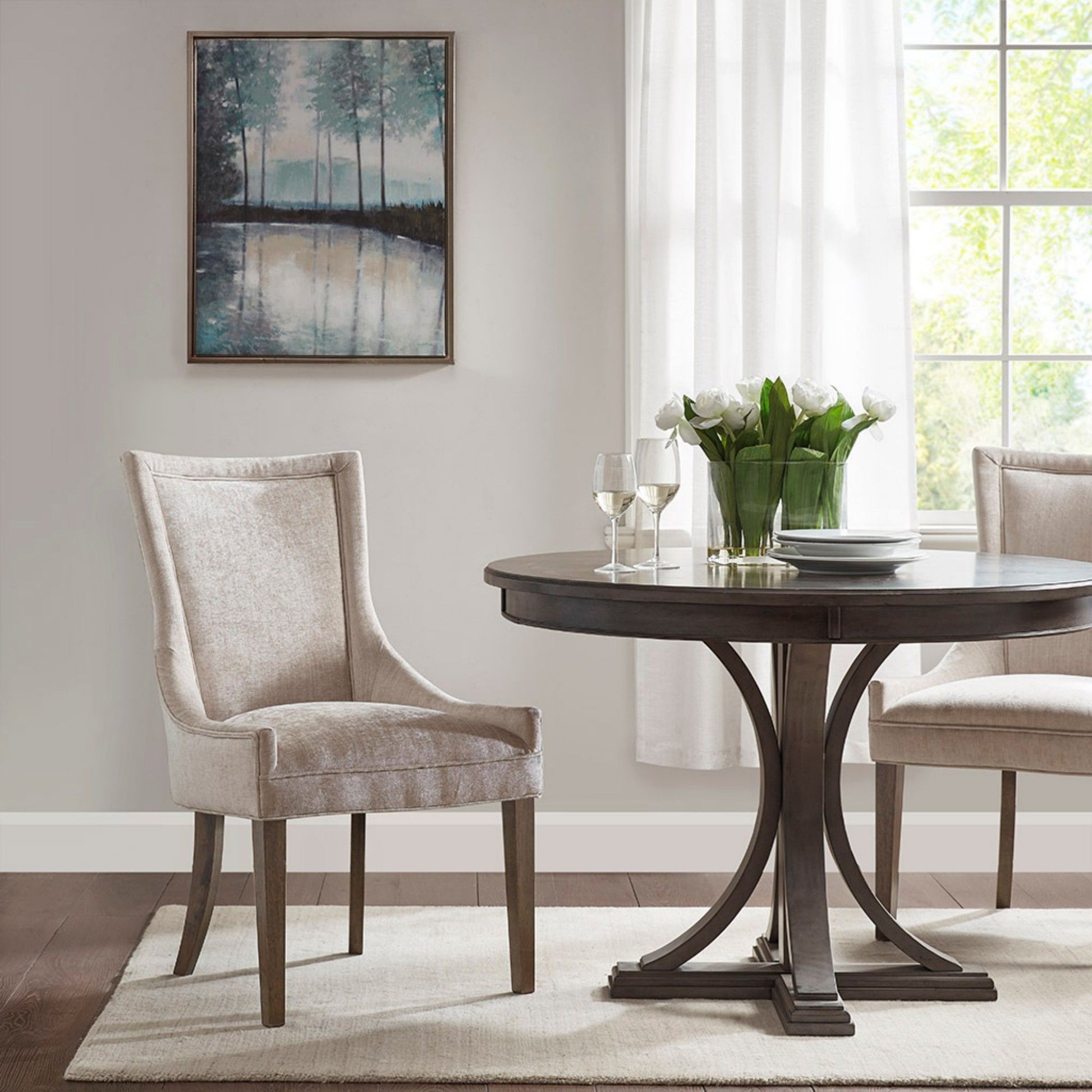 A round dark wood dining table with two Ultra - Elegant Dining Side Chairs in beige sits on a light rug. A vase of white tulips and two wine glasses are on the table. Sunlight streams in, and a landscape painting hangs on the wall.