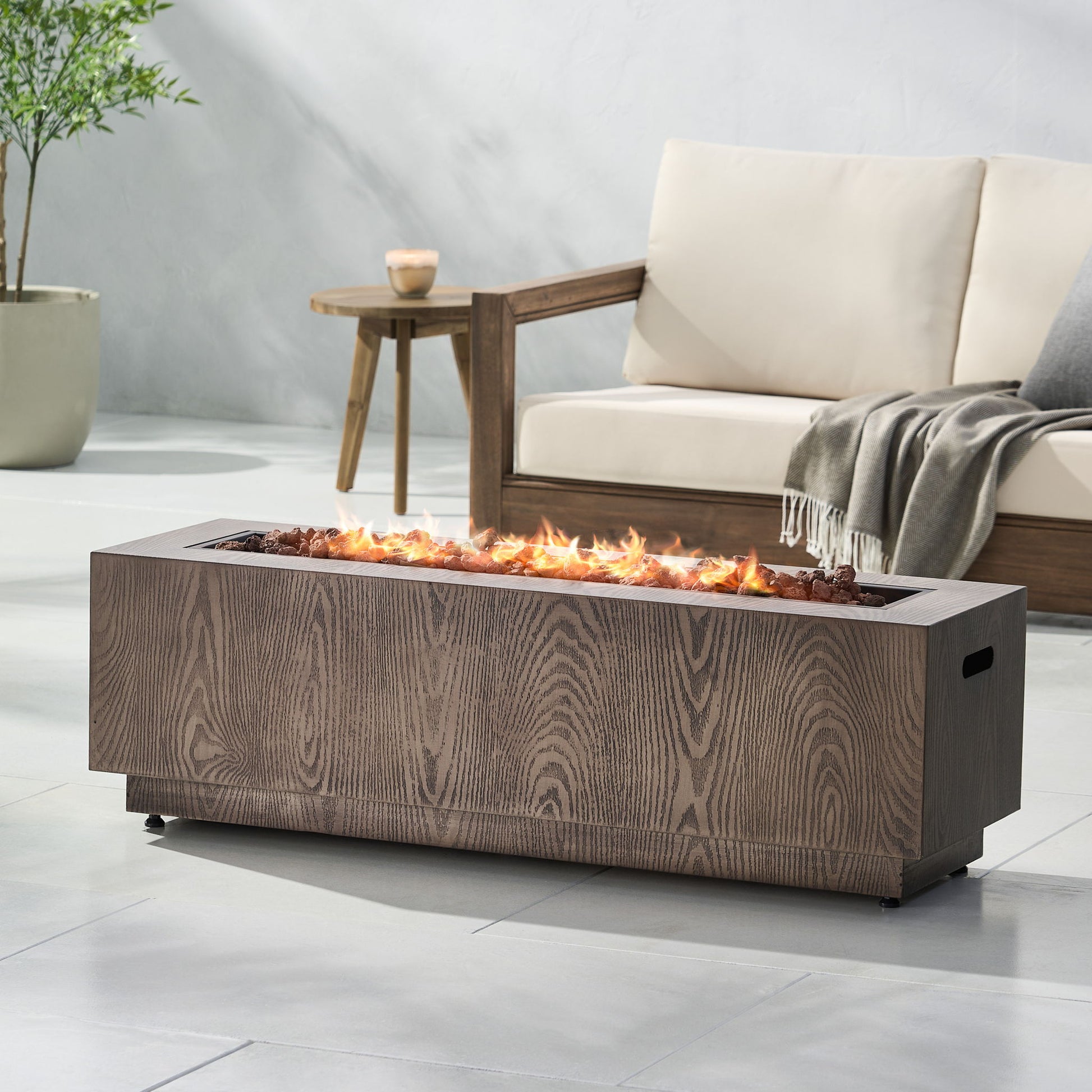 A modern patio with a beige cushioned sofa, wooden side table, potted plant, and the Wellington Square Iron Fire Pit With Wood Pattern Design and 50,000 Btu Output, set against a light gray wall and floor tiles.