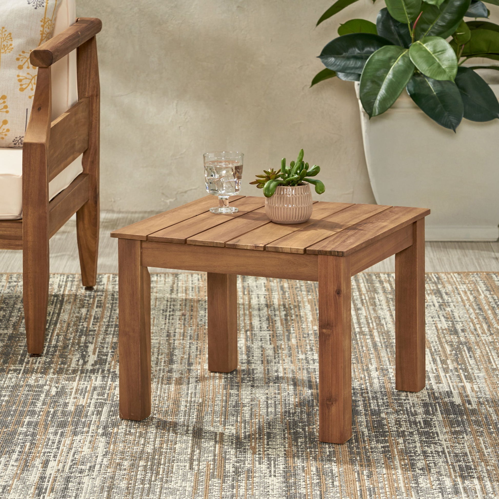 The Halcyon Outdoor Side Table With Slat Design, topped with a glass of water and a potted succulent, stands on a patterned rug beside a wooden chair, while a large potted plant decorates the background.