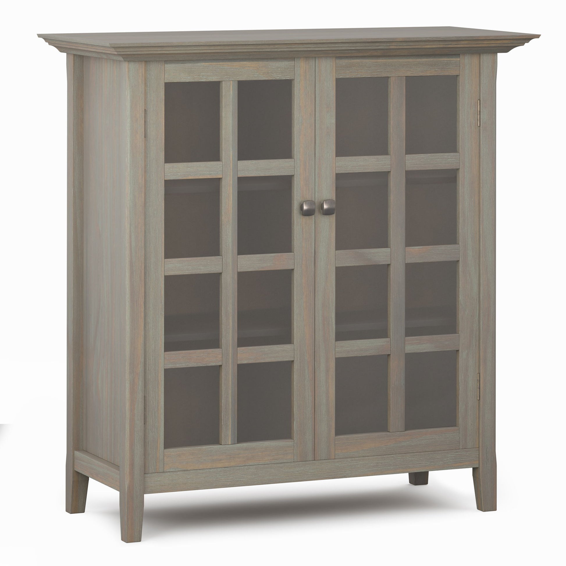 The Acadian - Medium Storage Cabinet is handcrafted with a light gray finish, two glass doors, metal knobs, three visible interior shelves, and stands on four short legs.
