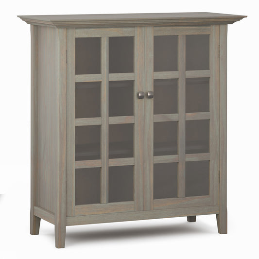 The Acadian - Medium Storage Cabinet is handcrafted with a light gray finish, two glass doors, metal knobs, three visible interior shelves, and stands on four short legs.
