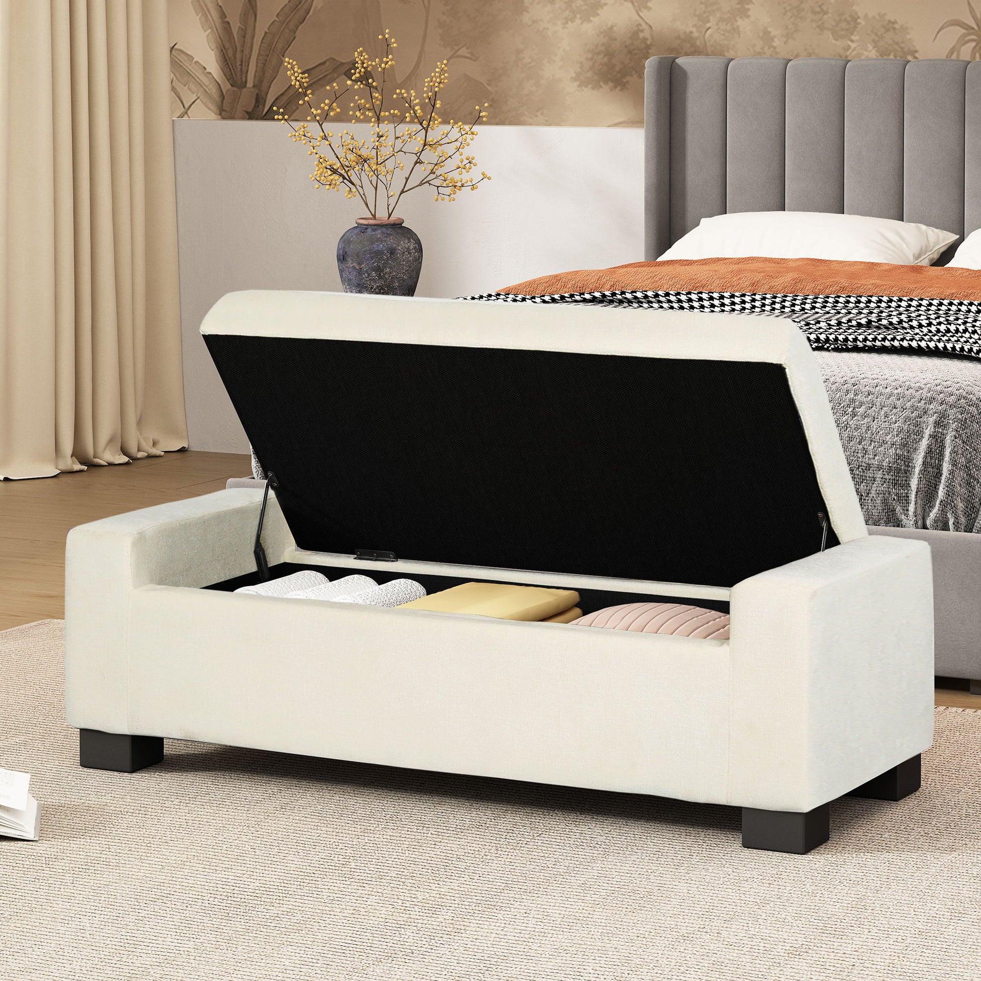 The Textured Fabric Ottoman Bench With Hinged Lid, shown open to reveal folded blankets and pillows inside, is placed at the foot of a bed in a modern, cozy bedroom.