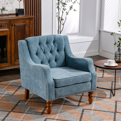 A button-tufted upholstered Accent Chair with vintage brass studs and footrest sits on a patterned rug beside a round wooden coffee table in a bright, stylish living room adorned with plants and wooden furniture.