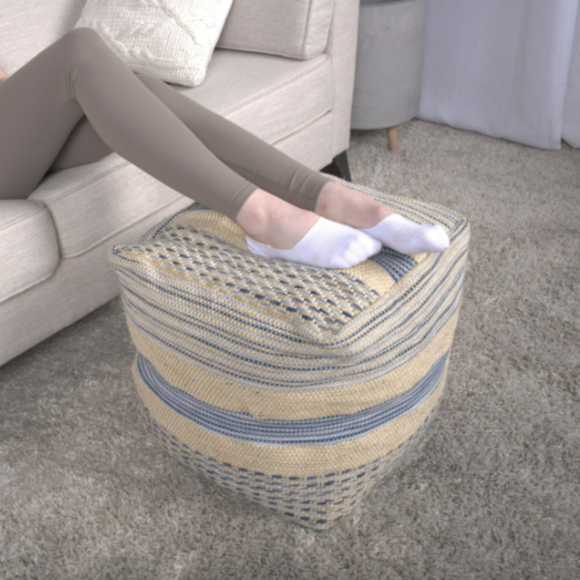 A person wearing white socks and beige leggings rests their feet on the Western - Handcrafted Fabric Pouf, Durable - Natural / Blue in a living room with a beige sofa and light gray carpet.