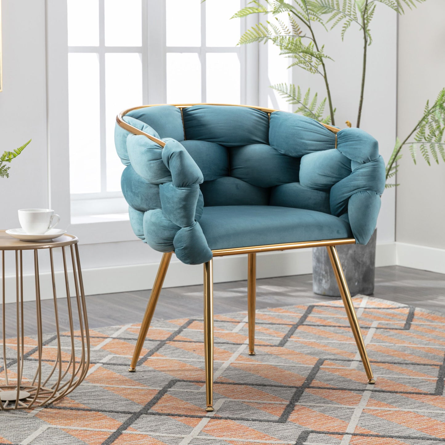 A Luxury Modern Simple Leisure Velvet Single Sofa Chair with plush teal upholstery and gold legs sits on a geometric rug; nearby are a round side table, teacup, and potted plant in a bright room.