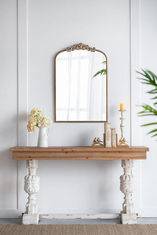 A wooden console table with ornate white legs stands against a white wall. On the table are a pitcher with flowers, books, a candlestick, and above hangs an Arch Mirror, Baroque Inspired Wall Decor reflecting light and greenery.