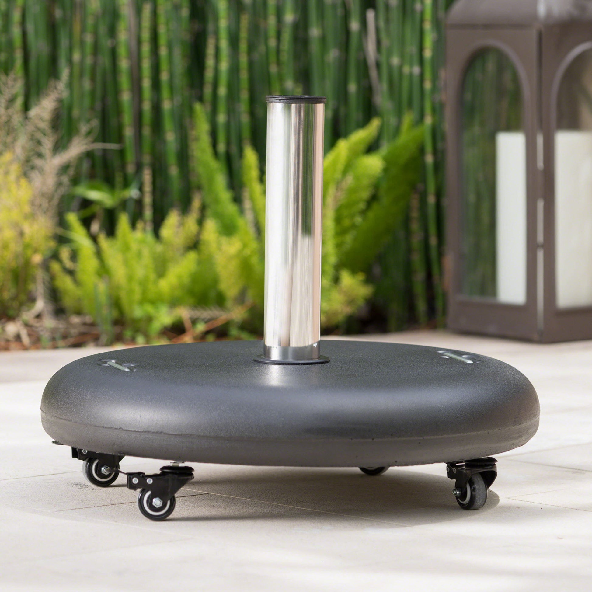 The Wheelie - Round Umbrella Base With Stainless Steel Holder features a black finish, silver holder, and four caster wheels. It sits on a patio with green plants and a tall lantern holding a white candle in the background.