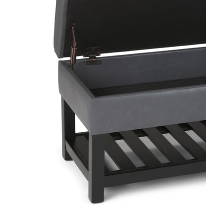 A close-up of the Cosmopolitan Storage Ottoman Bench With Open Bottom shows its black cushioned lid lifted open, revealing an empty storage compartment and a slatted shelf beneath, all supported by a dark wooden frame and legs.