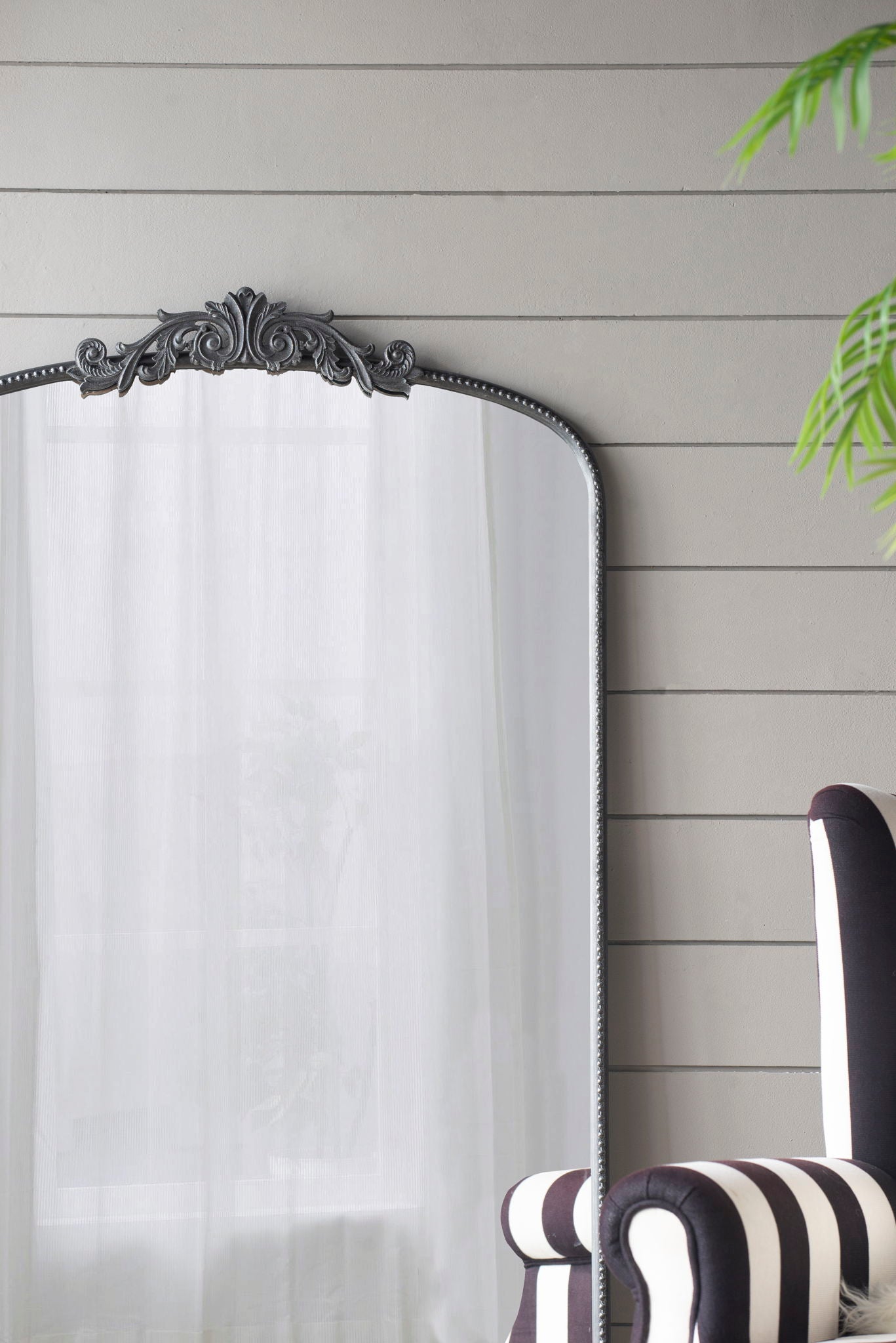 The Arch Mirror, a Baroque-inspired wall decor piece, stands against a gray shiplap wall reflecting a sheer white curtain. A black-and-white striped armchair and a green plant are visible to the right.