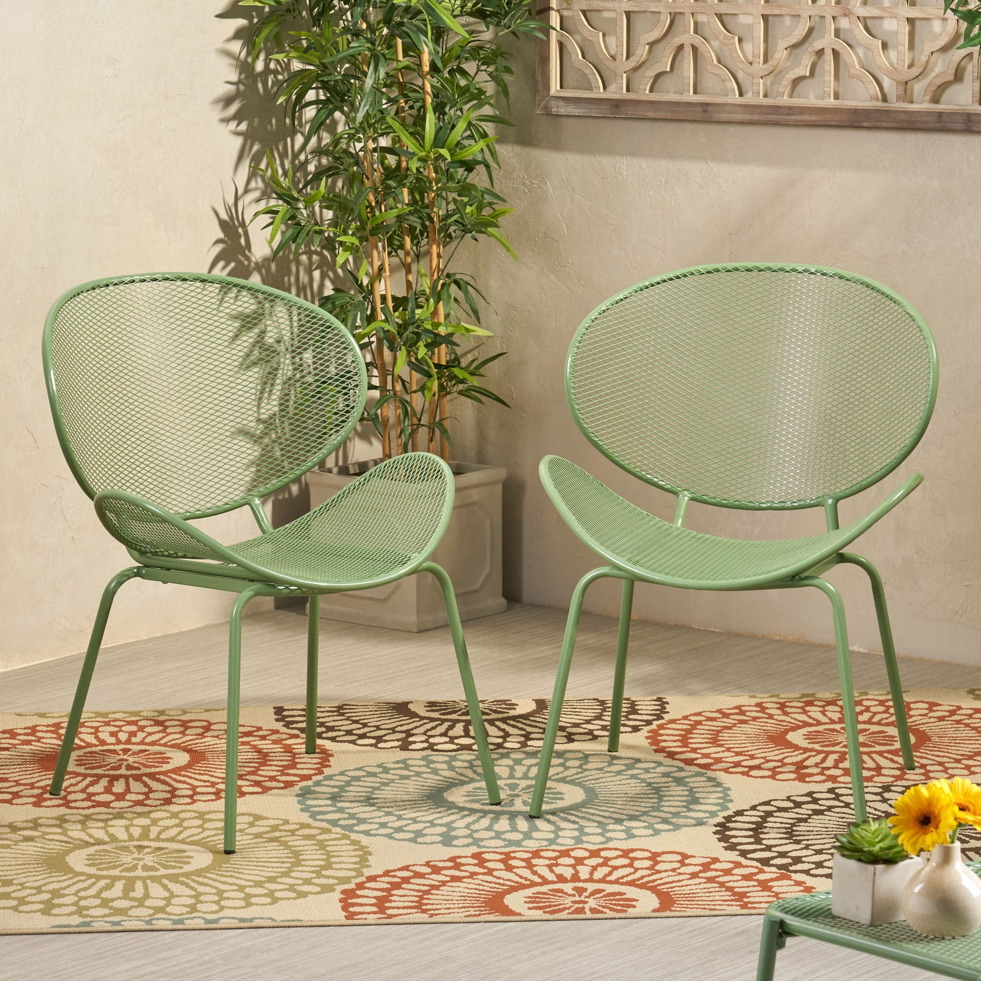 The Elba - Contemporary Chair Set For Outdoor Relaxation features two modern green metal chairs on a circular-patterned rug, with a potted plant, decorative wall panel in the background, and a small table with yellow flowers in front.