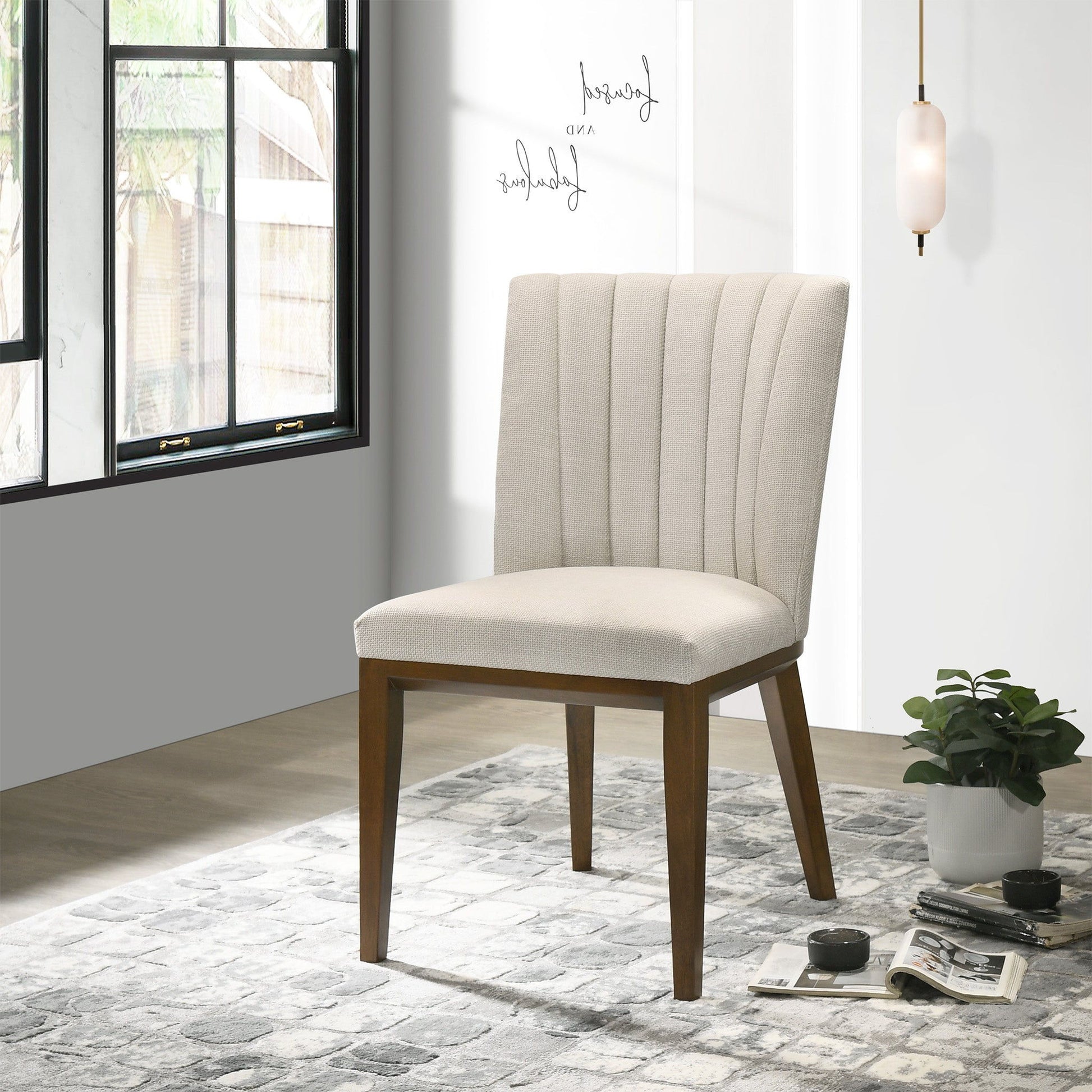 The Elmwood - Stylish Dining Chair (Set of 2), upholstered in modern beige with wooden legs, is placed on a patterned gray rug near a small plant, books, and teacup. Sunlight streams in while a pendant light hangs overhead.