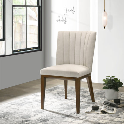 The Elmwood - Stylish Dining Chair (Set of 2), upholstered in modern beige with wooden legs, is placed on a patterned gray rug near a small plant, books, and teacup. Sunlight streams in while a pendant light hangs overhead.