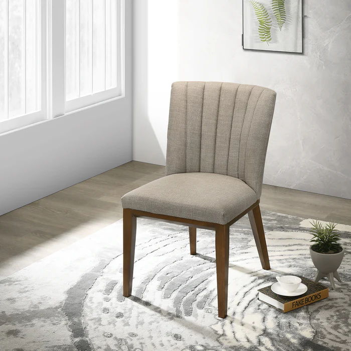 Two Elmwood Stylish Dining Chairs with wooden legs sit on a light patterned rug in a bright room. Nearby, a small table displays a potted plant, stacked books, and eyeglasses beneath a framed fern print on the wall.