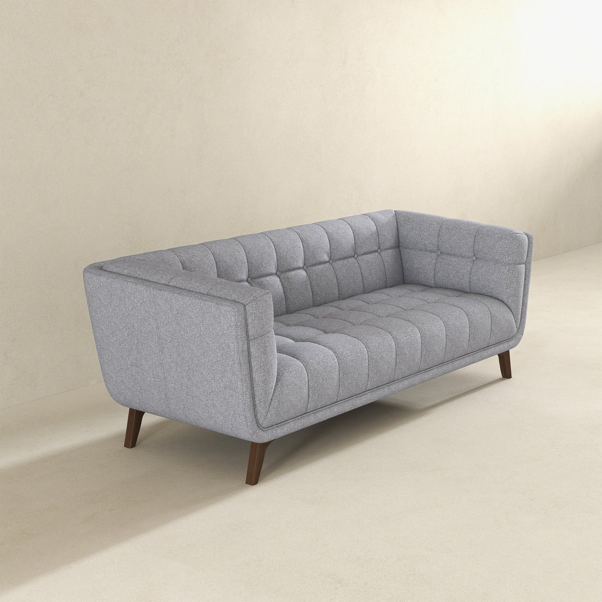 The Addison Mid-Century Modern Design Tufted Sofa, upholstered in gray with wooden legs, sits on a light beige floor against a plain, light-colored wall.