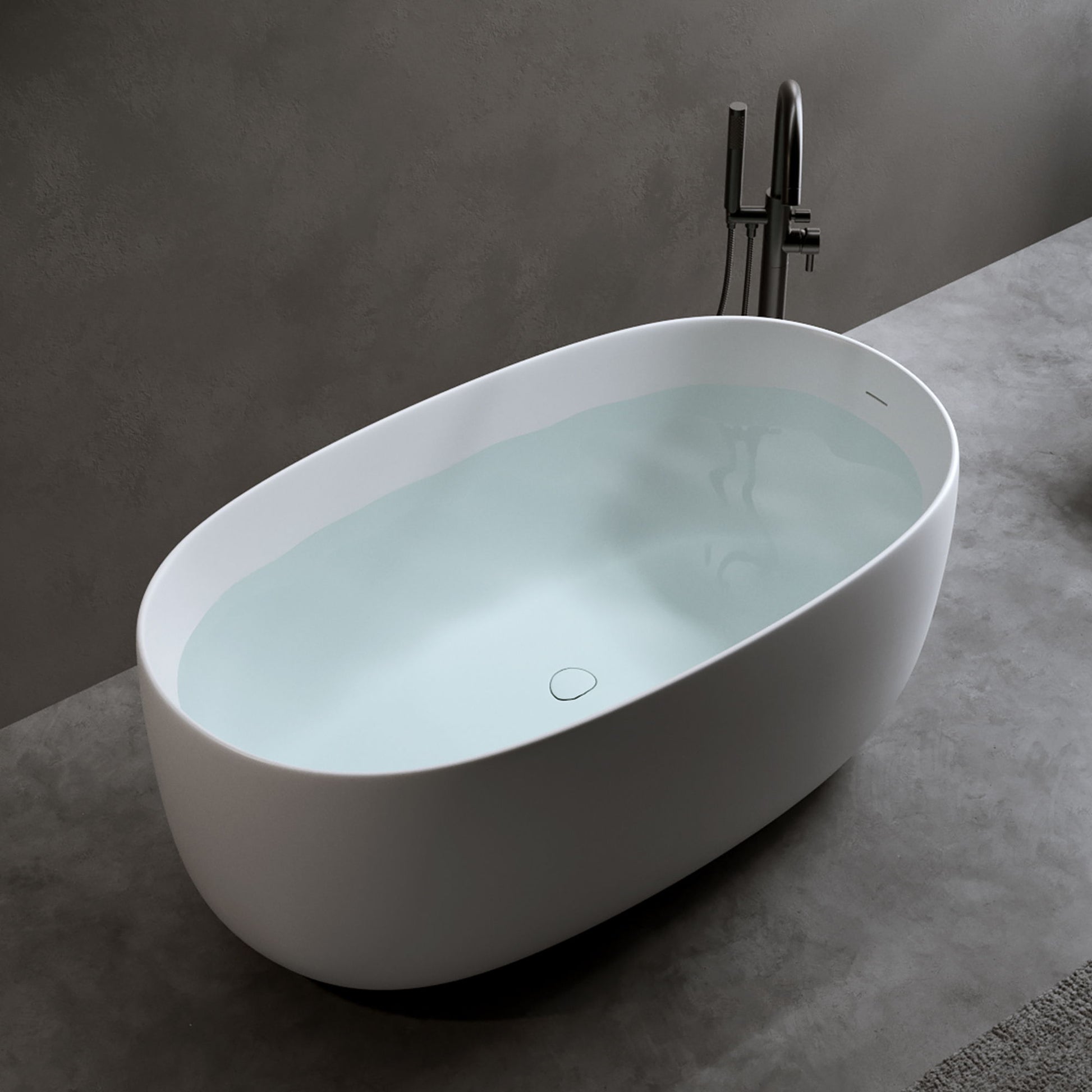 The Independent Solid Surface Resin Stone Bathtub in matte white, featuring a pop-up drain and overflow, is set against a gray wall and floor with a sleek tall faucet beside it—perfect for modern small households.