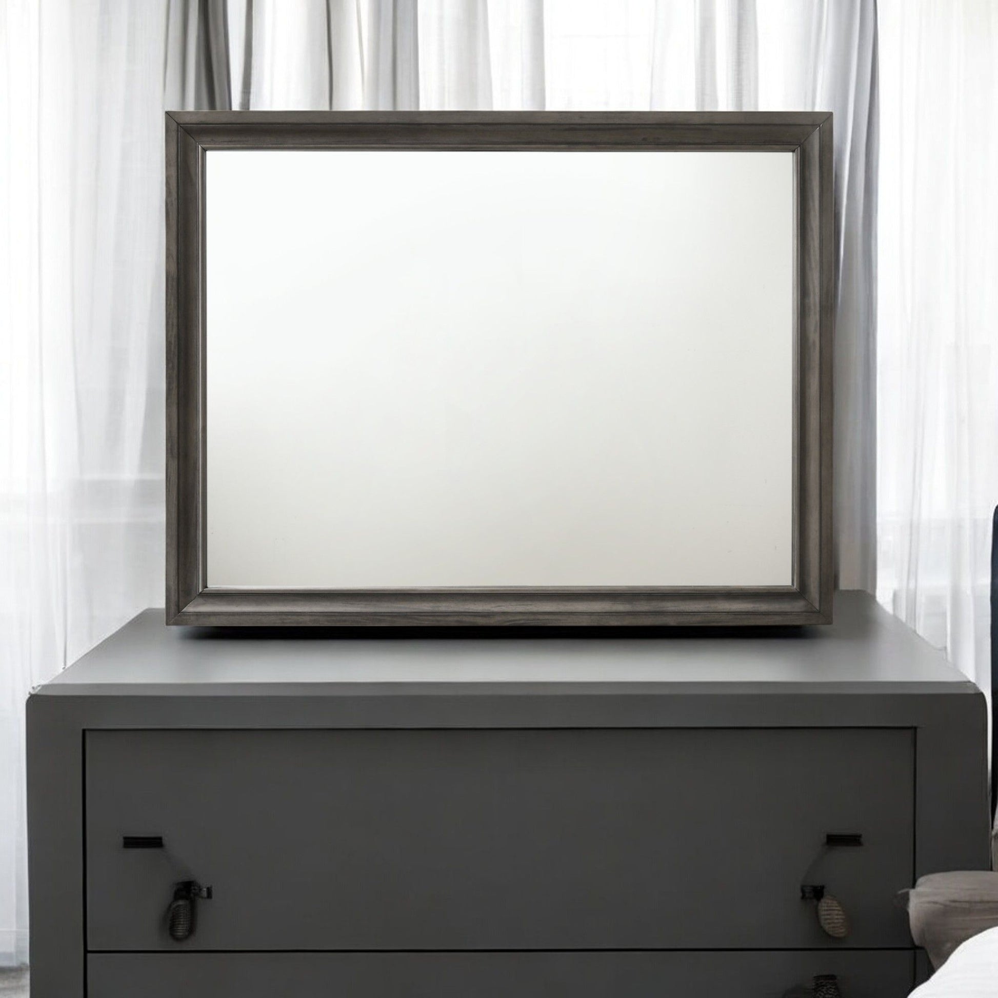 The Naima - Minimalist Design Mirror, featuring a large blank surface and a dark wooden frame, is placed atop a gray two-drawer dresser. It sits in front of sheer white curtains, enhancing the minimalist and clean setting.