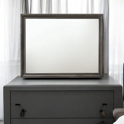 The Naima - Minimalist Design Mirror, featuring a large blank surface and a dark wooden frame, is placed atop a gray two-drawer dresser. It sits in front of sheer white curtains, enhancing the minimalist and clean setting.