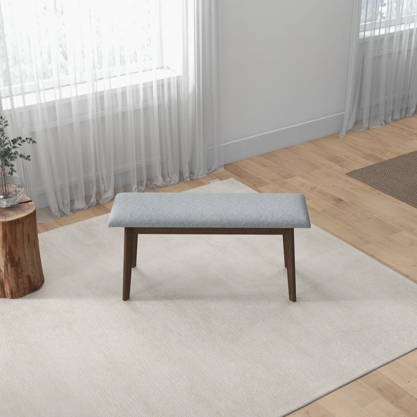 A minimalist room with light wood floors and white walls features the Carlos - Upholstered Bench in grey on a cream rug, paired with a small stump table holding a plant to the left and sheer curtains at the window.