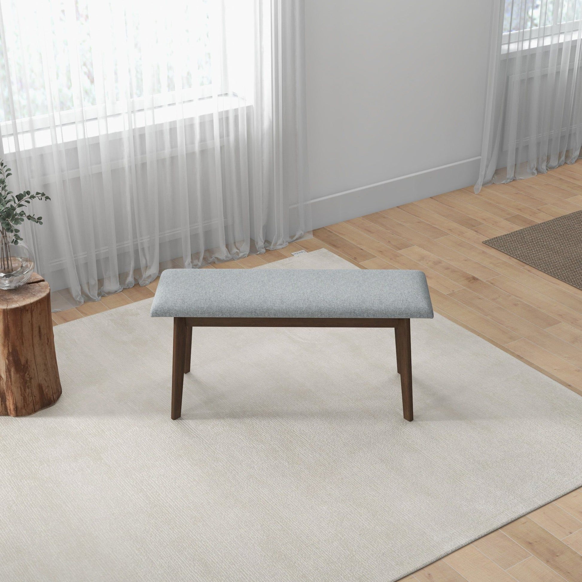 A minimalist room with light wood floors and white walls features the Carlos - Upholstered Bench in grey on a cream rug, paired with a small stump table holding a plant to the left and sheer curtains at the window.