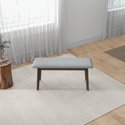 A minimalist room with light wood floors and white walls features the Carlos - Upholstered Bench in grey on a cream rug, paired with a small stump table holding a plant to the left and sheer curtains at the window.
