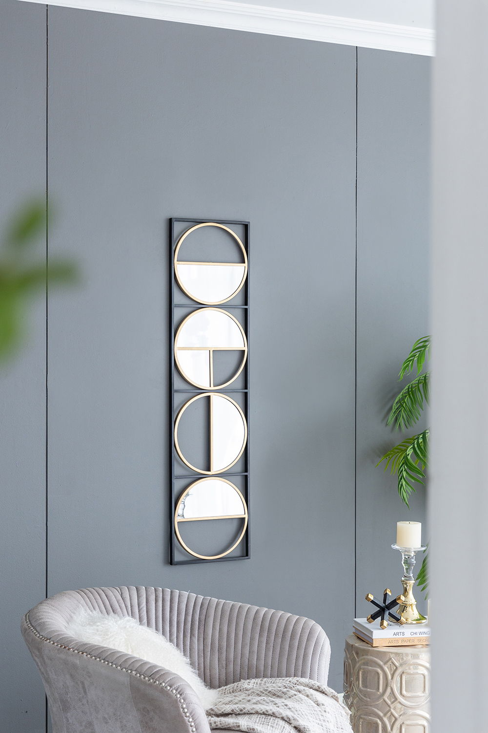 A modern living room with gray walls showcases the Eclectic Styling Metal Beaded Wall Mirror, featuring four gold-framed circular mirrors. Nearby are a plush gray chair, a small candlelit table, and a leafy plant for decoration.