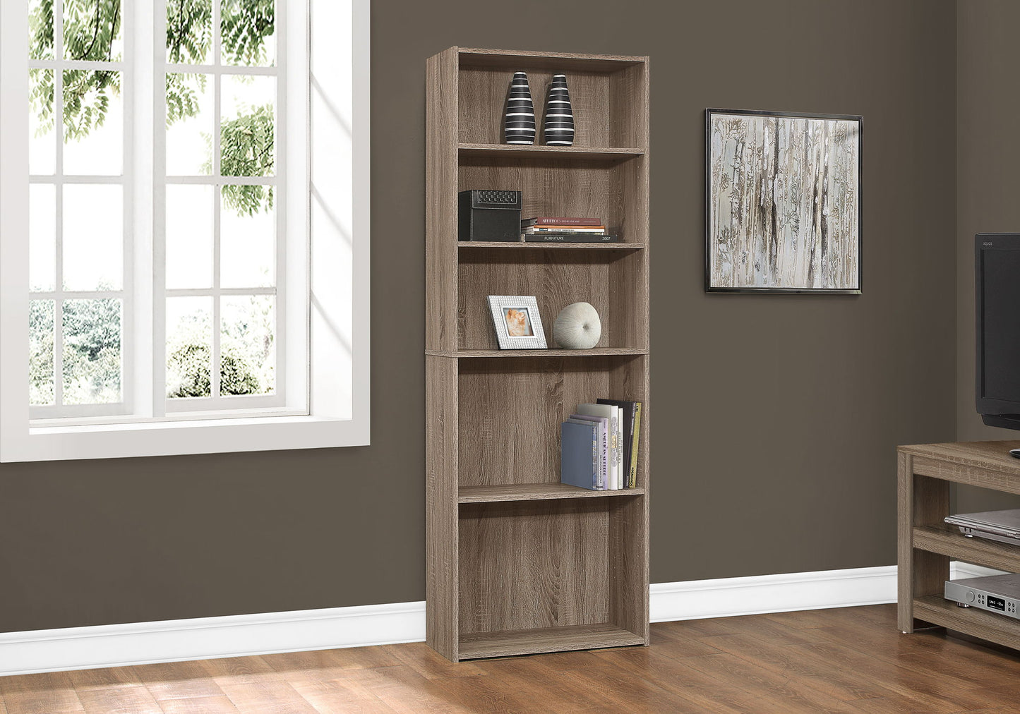 A 6-tier transitional bookshelf stands against a brown wall in the living room with wooden floors, a window, framed art, and a TV stand, displaying vases, books, and decor.